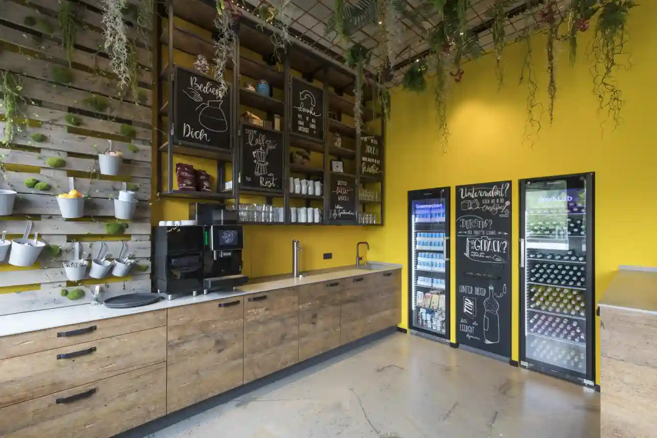 Gewerbliche Küche mit Holzschränken, Kaffeeautomat, Hängeregale, Kühlschränken und gelber Wand.