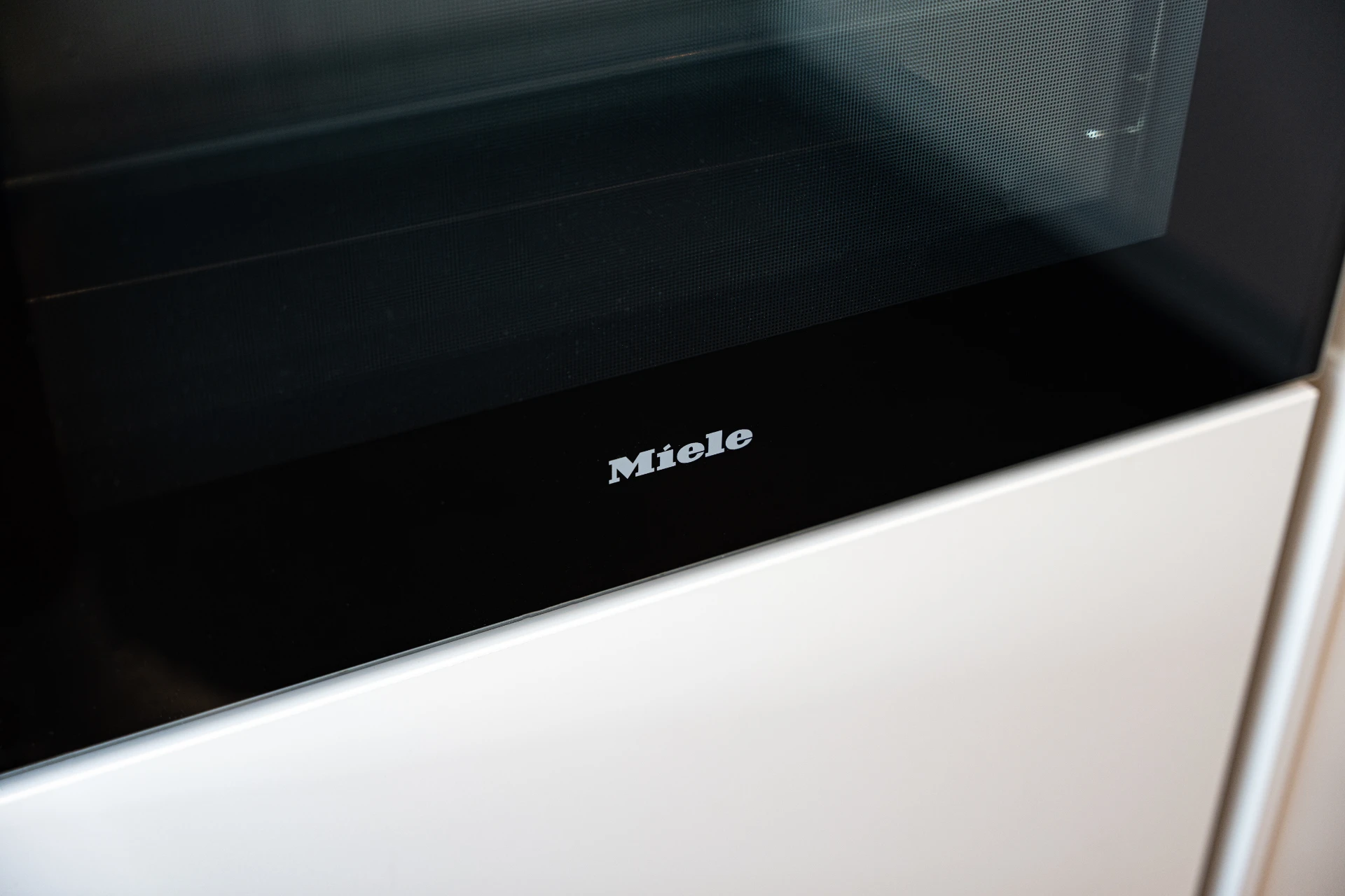 Naheaufnahme des Miele-Logos auf einem schwarzen Ofenpanel mit weißer Ofentür darunter.