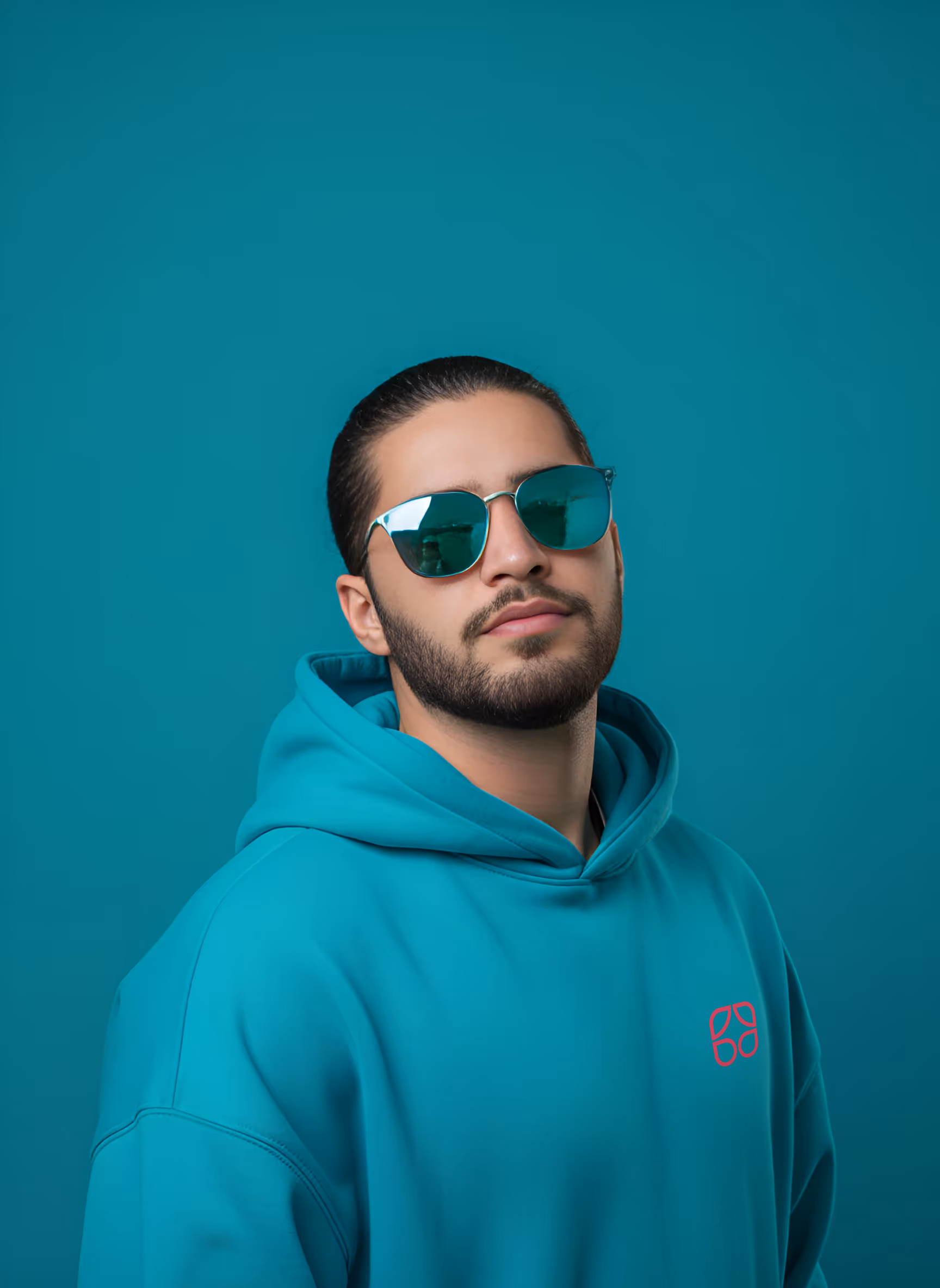გრძელ თმიანი, წვერიანი მამაკაცი ტურკუაზისფრო სპორტულ კოსტიუმსა და სათვალეს აცვია ტურკუაზისფერ ფონზე.