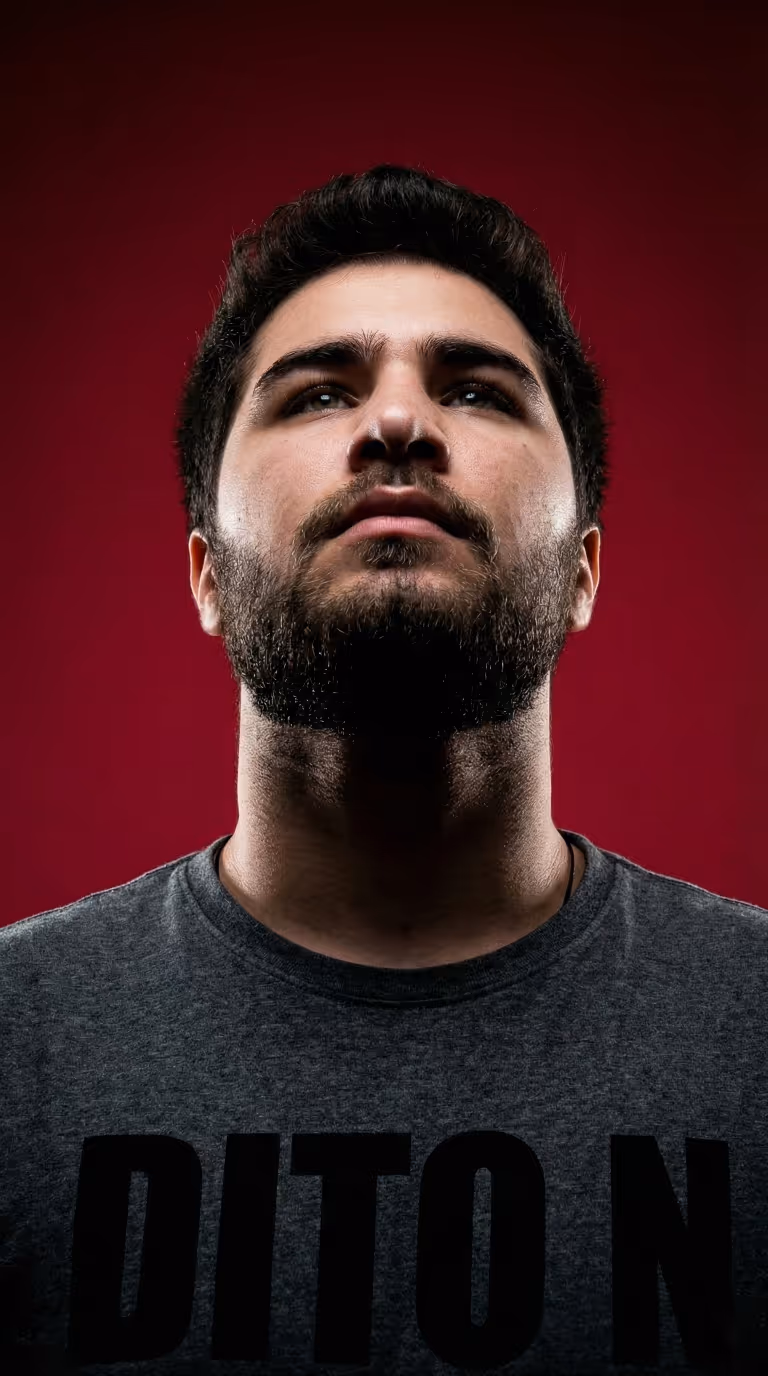 ძალიან გრძელი მაისურიანი მამაკაცი, რომელიც აფართხალებული მუქი წვერით და შავი თმით, უზარმაზარი წითელი ფონის წინ იყურება მაღლა.