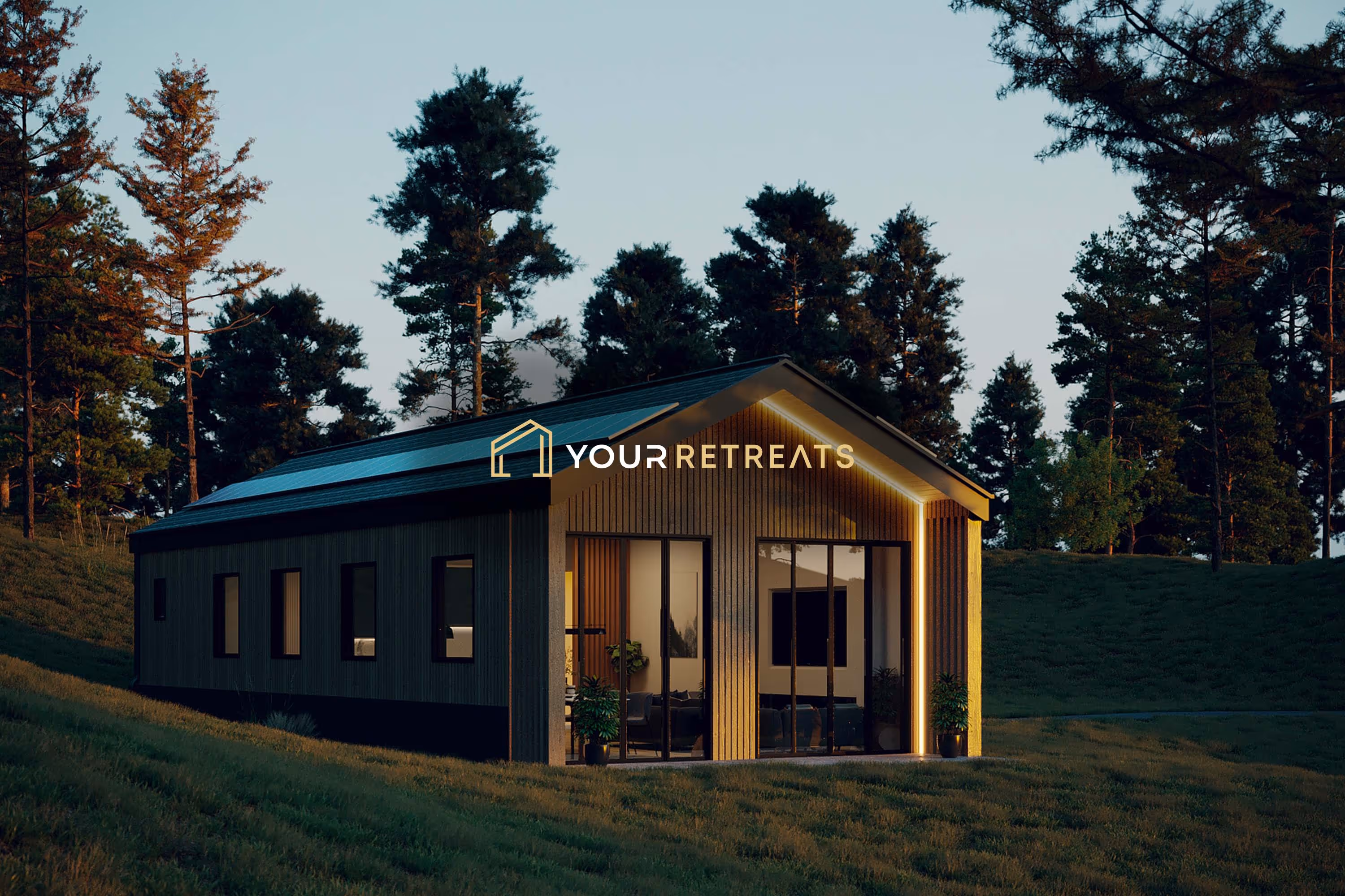 Modern cabin with large glass doors lit warmly, set in a grassy, tree-surrounded landscape at dusk.