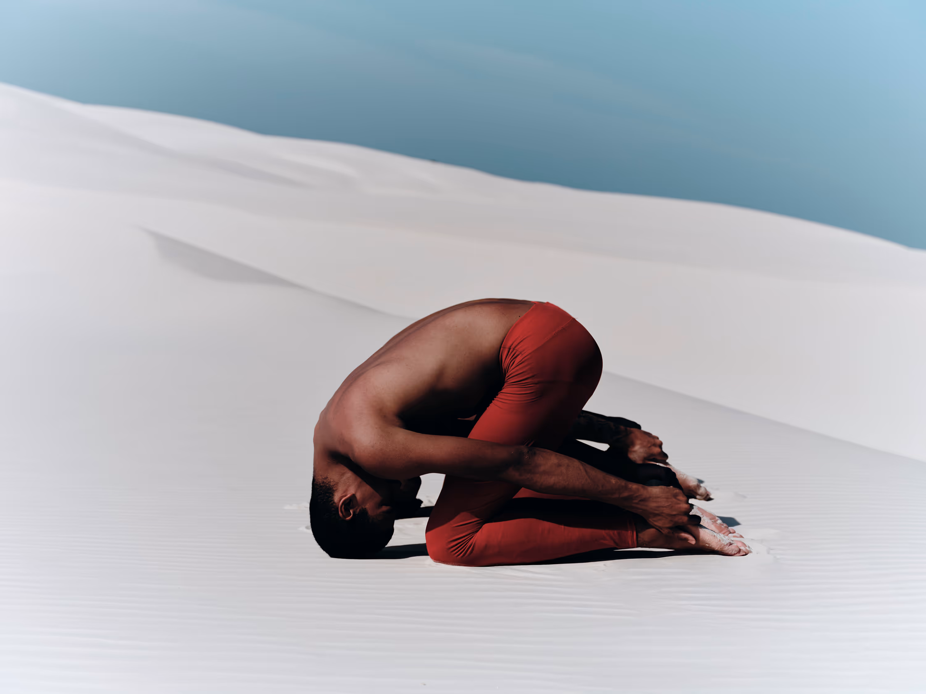 Man wearing red pants performing a yoga child's pose on white sand dunes under a clear blue sky.