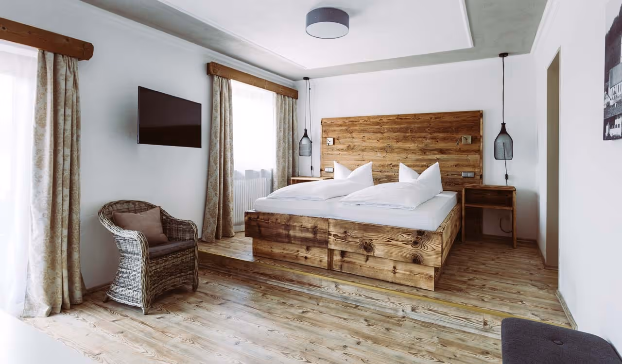 Bright bedroom with wooden platform bed, wicker chair, bedside tables, hanging lamps, and a wall-mounted TV.