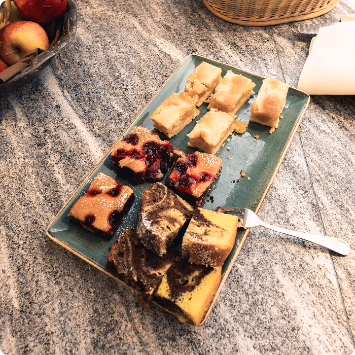 Rectangular plate with a variety of sliced cakes including berry, vanilla, and marbled chocolate pieces on a granite countertop next to a basket of apples and a fork.