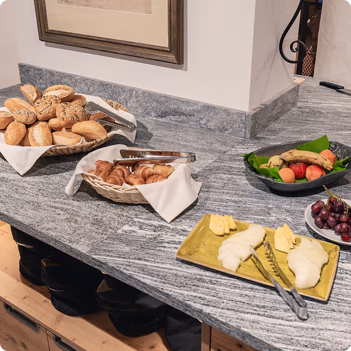 Granite countertop with baskets of assorted bread and croissants, a rectangular plate with sliced cheese, and a bowl of fruit including grapes, apples, and bananas.