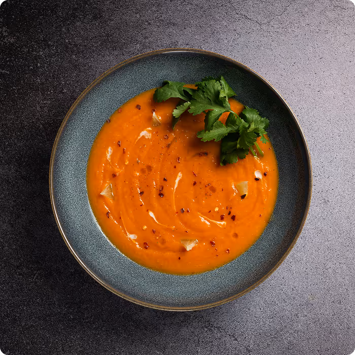 Bowl of creamy orange soup garnished with fresh cilantro, chili flakes, and garlic slices on a dark textured surface.