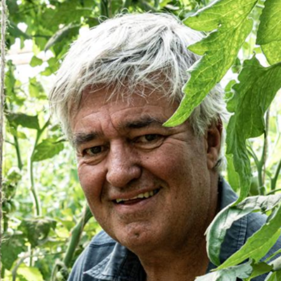 Smiling older man with gray hair surrounded by green leaves in a garden.