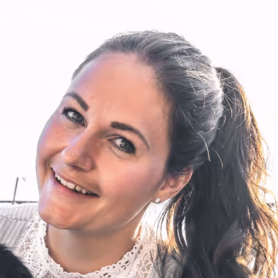 Smiling woman with dark hair tied back, wearing a lace-collared top and sweater, against a bright background.