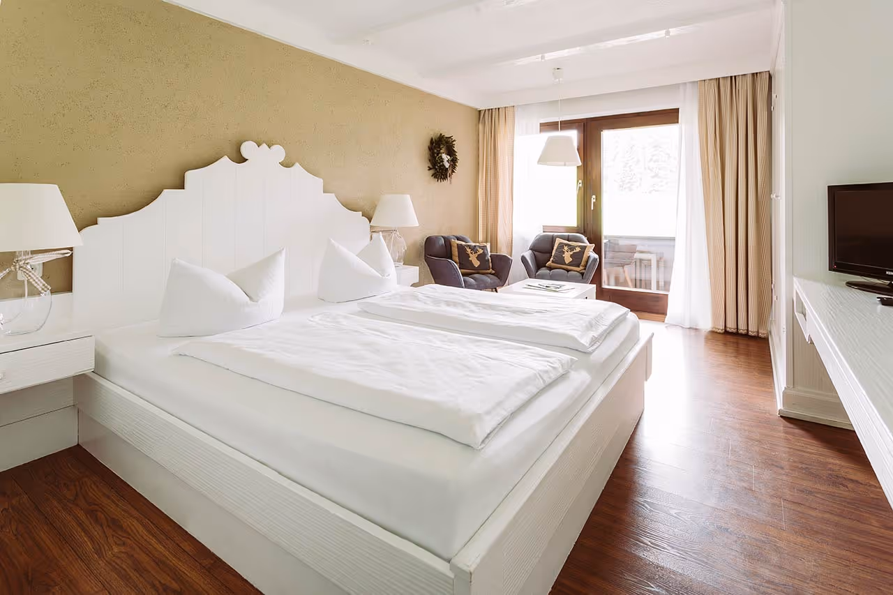 Bright hotel room with white double bed, two armchairs with deer cushions, wooden floor, and a window with curtains.
