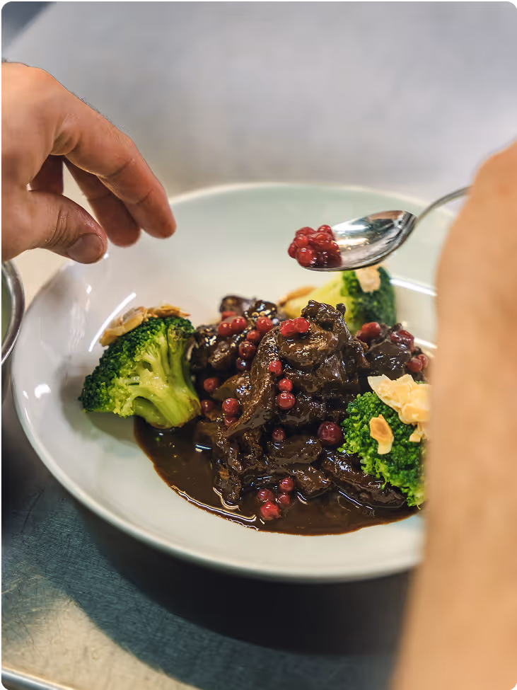 Close-up of a white plate with cooked beef in dark sauce, garnished with red berries, broccoli florets, and almond slices, with a hand adding berries with a spoon.