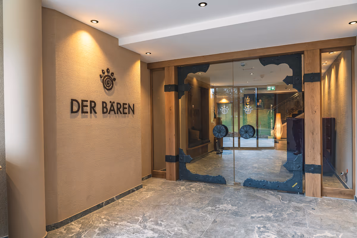 Entrance hallway with beige walls, the words 'DER BÄREN' on the left wall, and glass doors with decorative black metal and wooden frame.