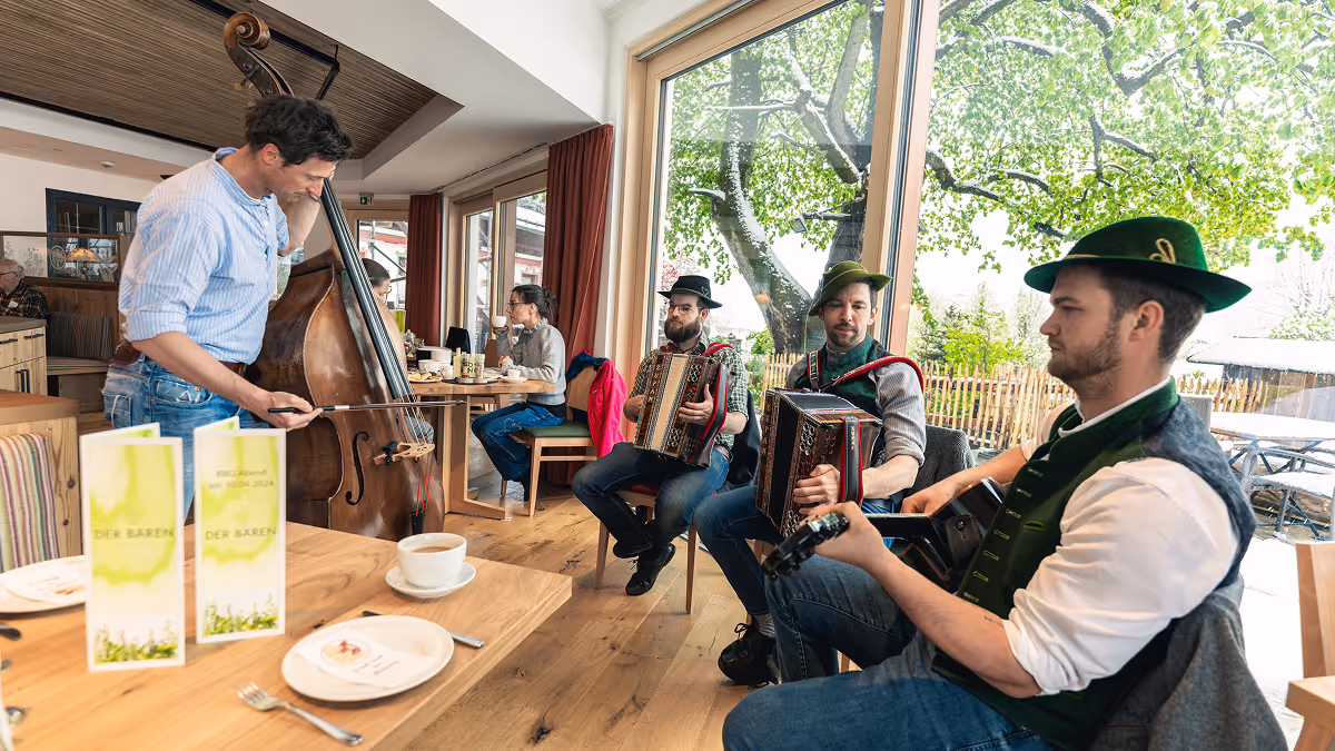 Four musicians in traditional attire playing guitar, accordion, and double bass inside a cozy café with large windows showing green trees outside.