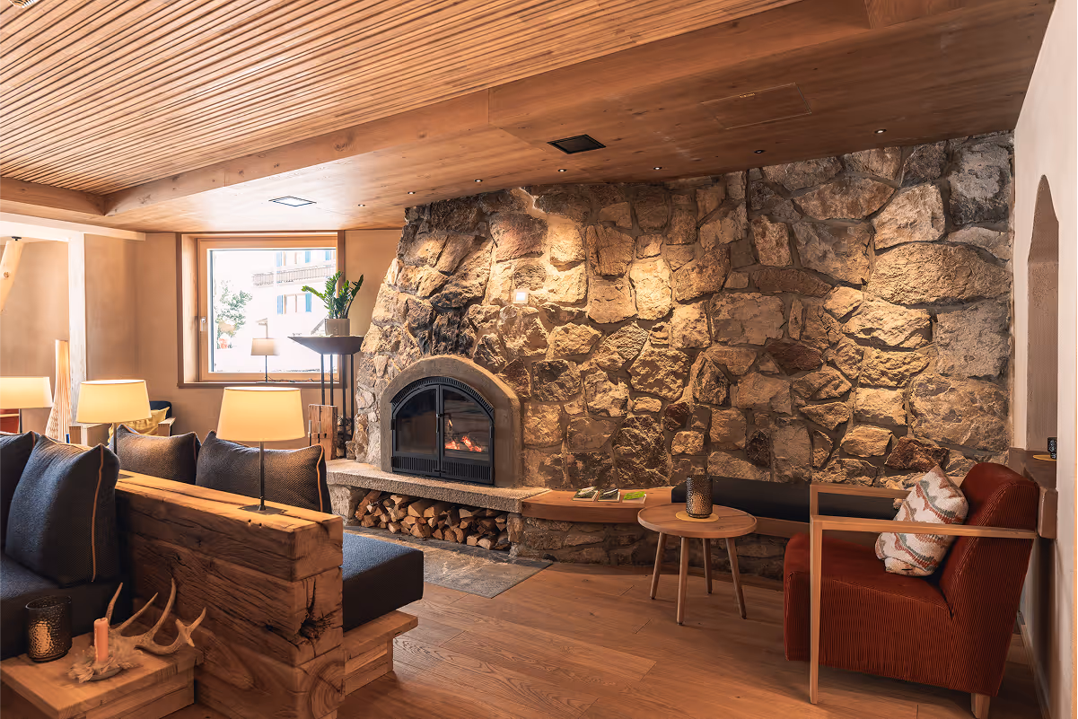 Cozy living room with a stone fireplace, wooden ceiling and floor, a dark cushioned sofa, a red armchair, and warm lighting.