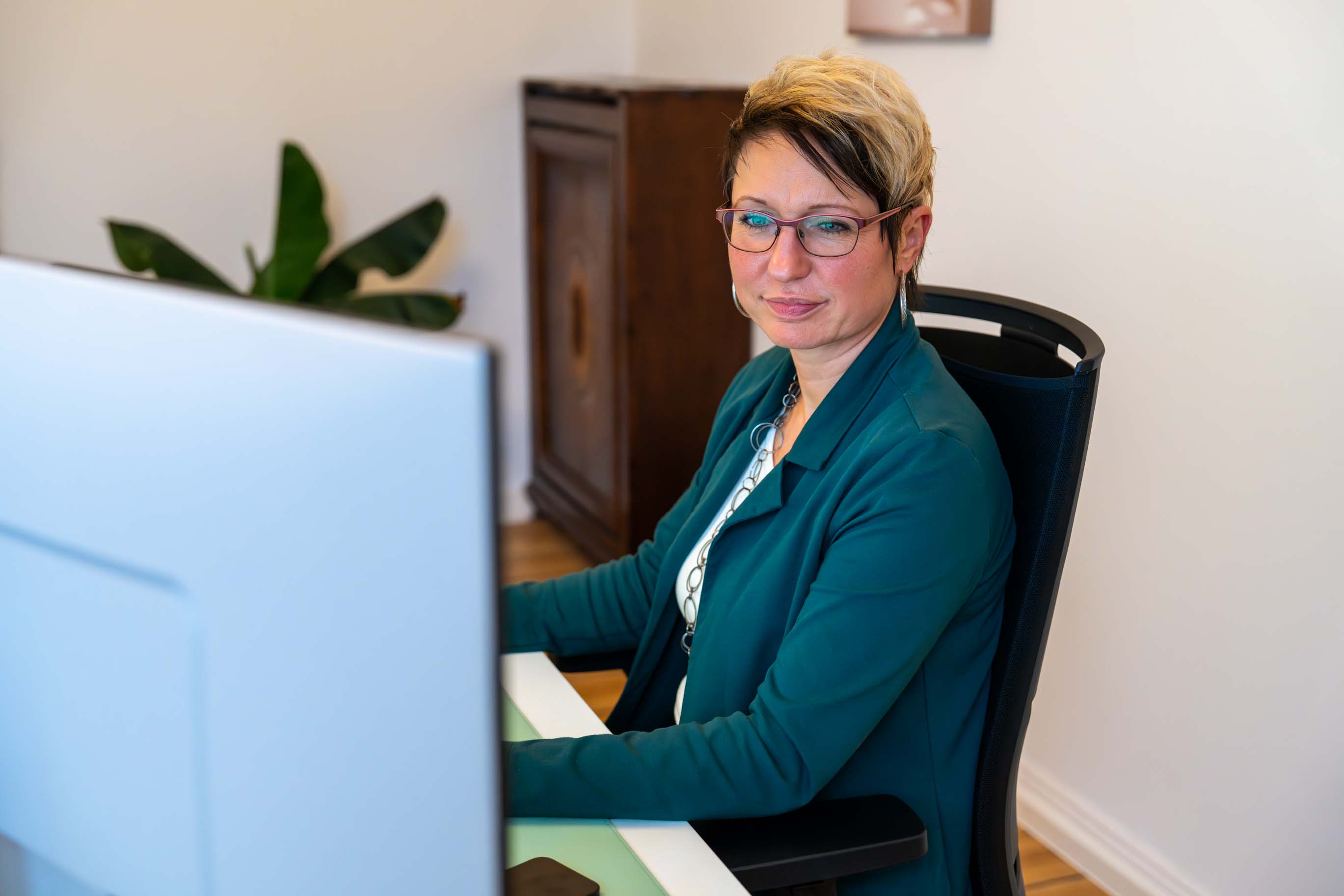 Mitarbeiterin mit kurzen blond-braunen Haaren, Brille und einem blaugrünen Blazer, die in einem Büro am Computer arbeitet.