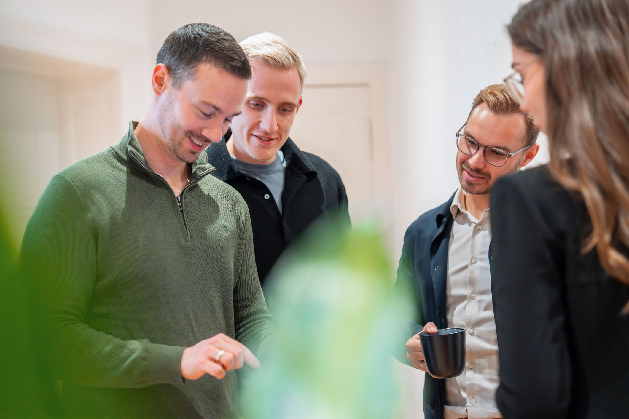 Vier Kollegen stehen zusammen in einem Büro, zwei Männer schauen auf ein Tablet, ein Mann hält eine schwarze Tasse in der Hand und eine Frau steht ihnen gegenüber.