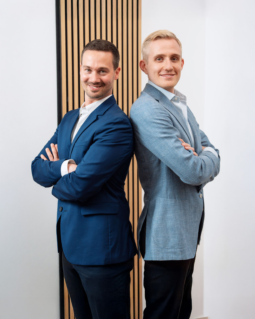 Zwei lächelnde Geschäftsführer stehen Rücken an Rücken mit verschränkten Armen und tragen Anzüge.