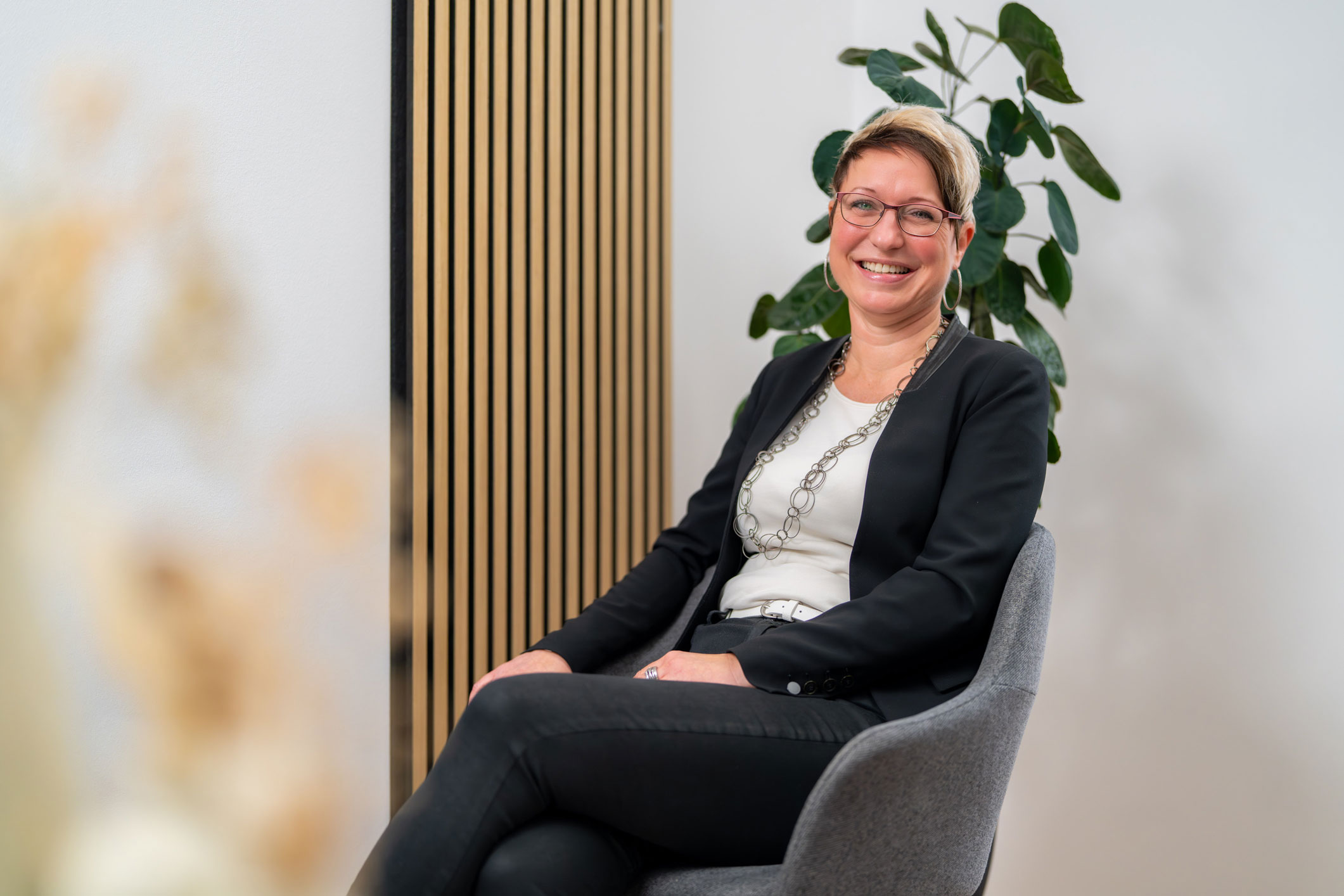 Lächelnde Frau mit Brille und kurzen blonden Haaren, die in einem modernen Büro auf einem grauen Stuhl sitzt. Hinter ihr steht eine Pflanze.