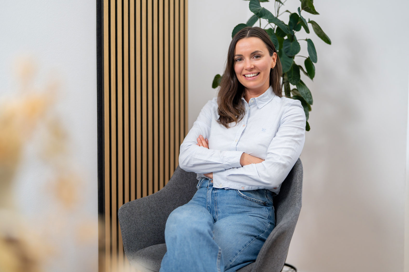 Lächelnde Frau mit braunen Haaren, die ein hellblaues Hemd und Jeans trägt und mit verschränkten Armen auf einem grauen Stuhl sitzt, in einem Innenraum mit einer Pflanze und einer Holzvertäfelung im Hintergrund.