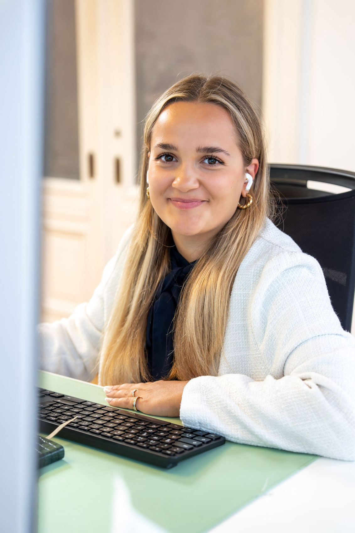 Lächelnde Mitarbeiterin mit langen blonden Haaren, die einen weißen Pullover und kabellose Ohrhörer trägt, sitzt an einem Schreibtisch mit einer Tastatur.