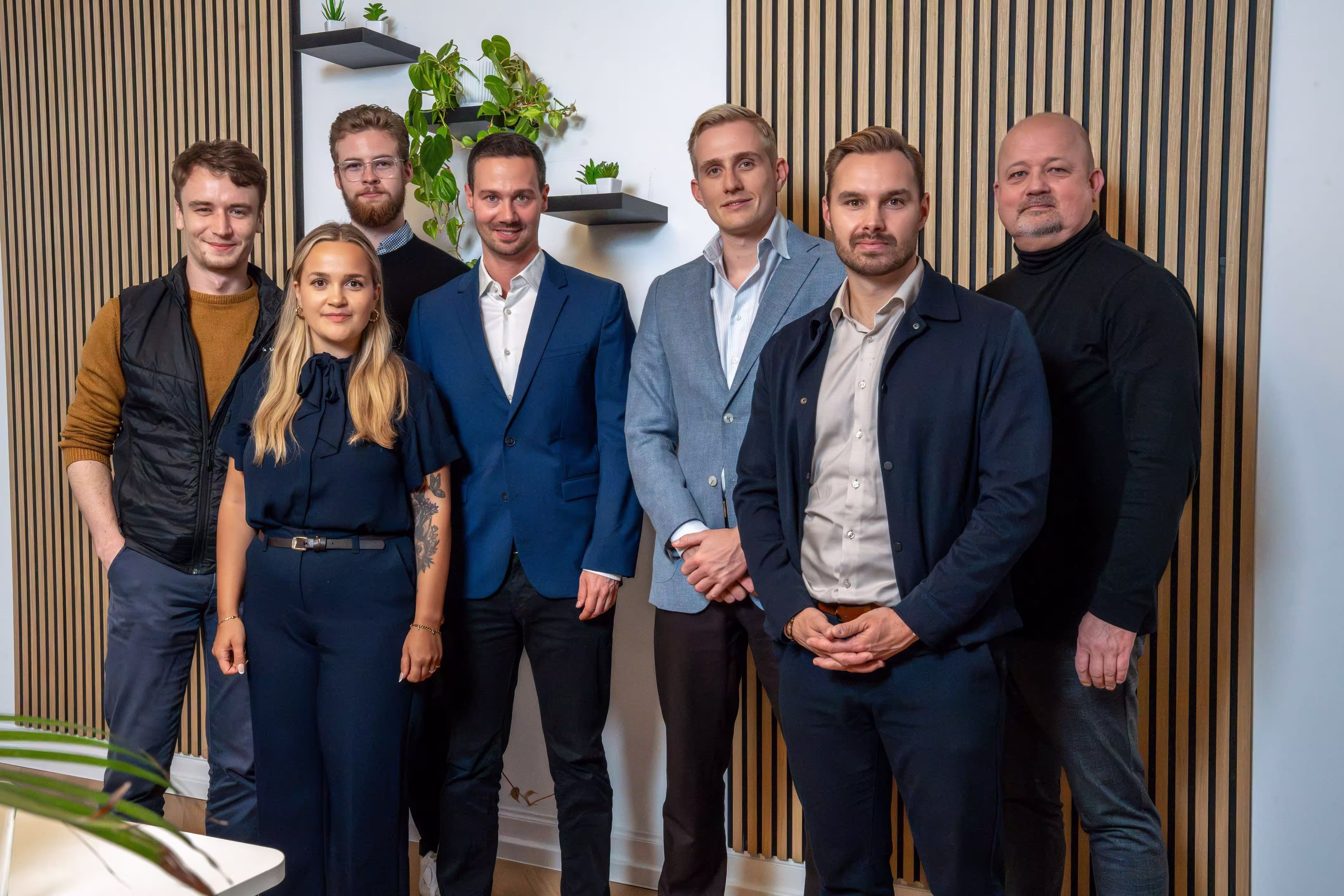 Gruppe von sieben Fachleuten, die in einem Innenraum vor einer holzgetäfelten Wand stehen, an der kleine Wandregale mit Pflanzen angebracht sind.