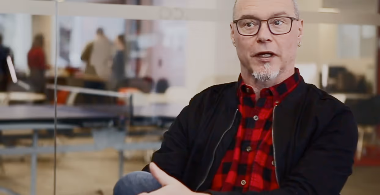 Middle-aged man with glasses and a goatee wearing a red and black checkered shirt speaking in a modern office space.