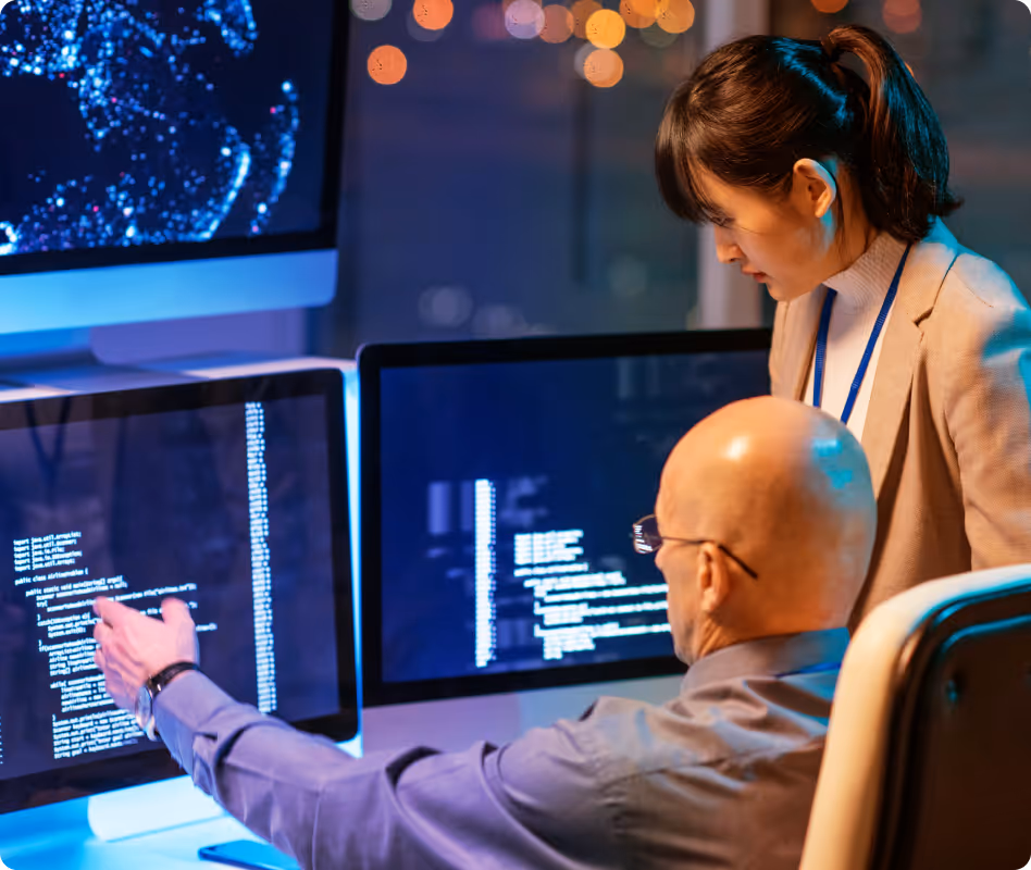 Two professionals analyzing computer code on multiple monitors in a dimly lit office.