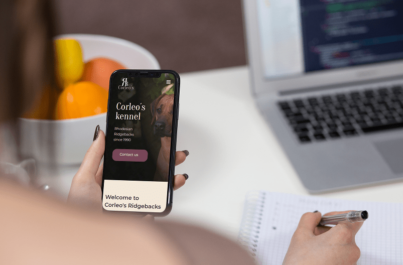 Person holding a smartphone showing Corleo's Kennel website with a Rhodesian Ridgeback dog image, beside a laptop and a notebook with a pen.