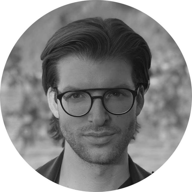 Portrait of a man with medium-length hair, wearing round glasses and light facial hair, looking into the camera with a blurred background.