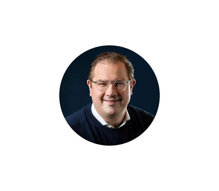 Lächelnder Mann mit heller Haut, kurzem braunem Haar, der eine klare Brille, ein weißes Hemd mit Kragen und einen dunklen Pullover trägt, vor dunkelblauem Hintergrund.