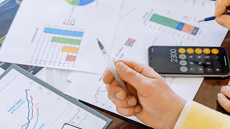 Zwei Personen analysieren Finanzdiagramme auf Papier und Tablet, eine Person hält einen Stift, ein Taschenrechner zeigt 23,00.