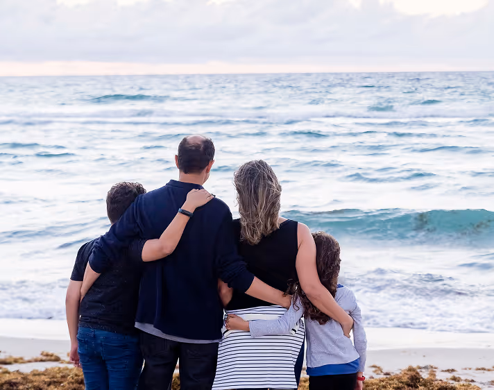 Familie mit zwei Erwachsenen und zwei Kindern umarmt sich am Strand und blickt auf das Meer.