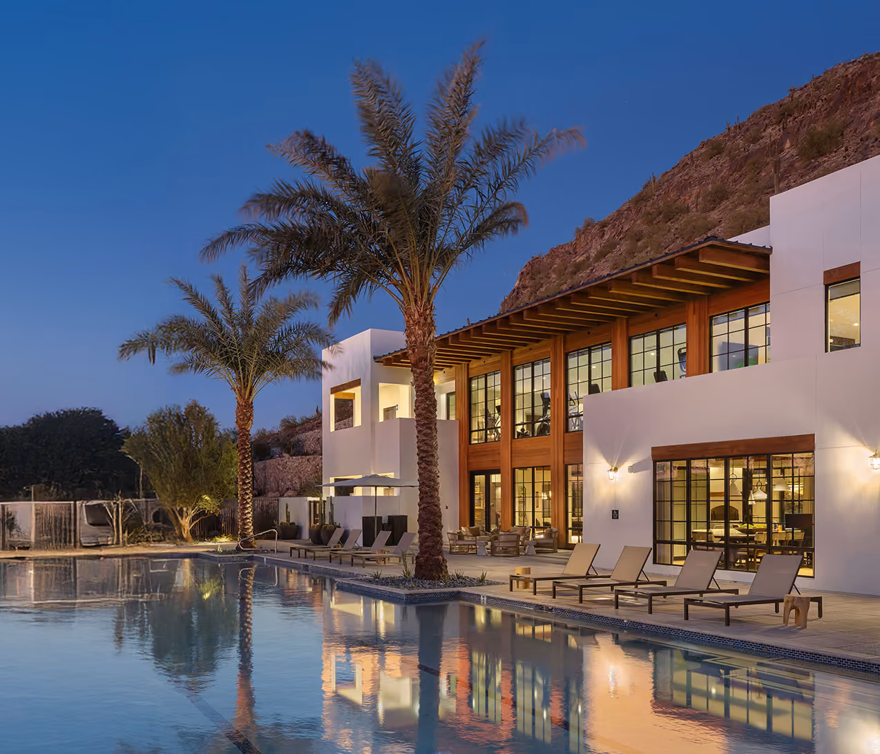Modern house with large windows, palm trees, and a pool reflecting the building at dusk.