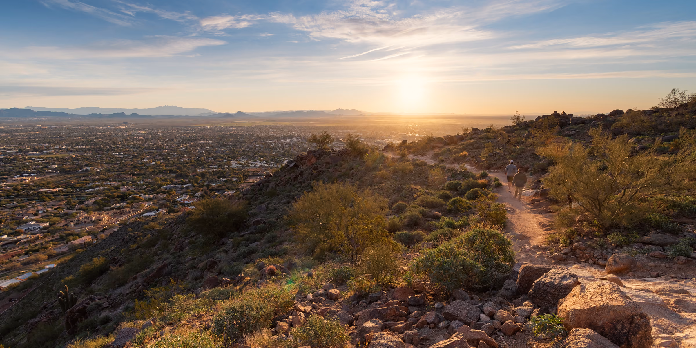 Two people hiking on a rocky trail overlooking a city at sunset with mountains in the distance.