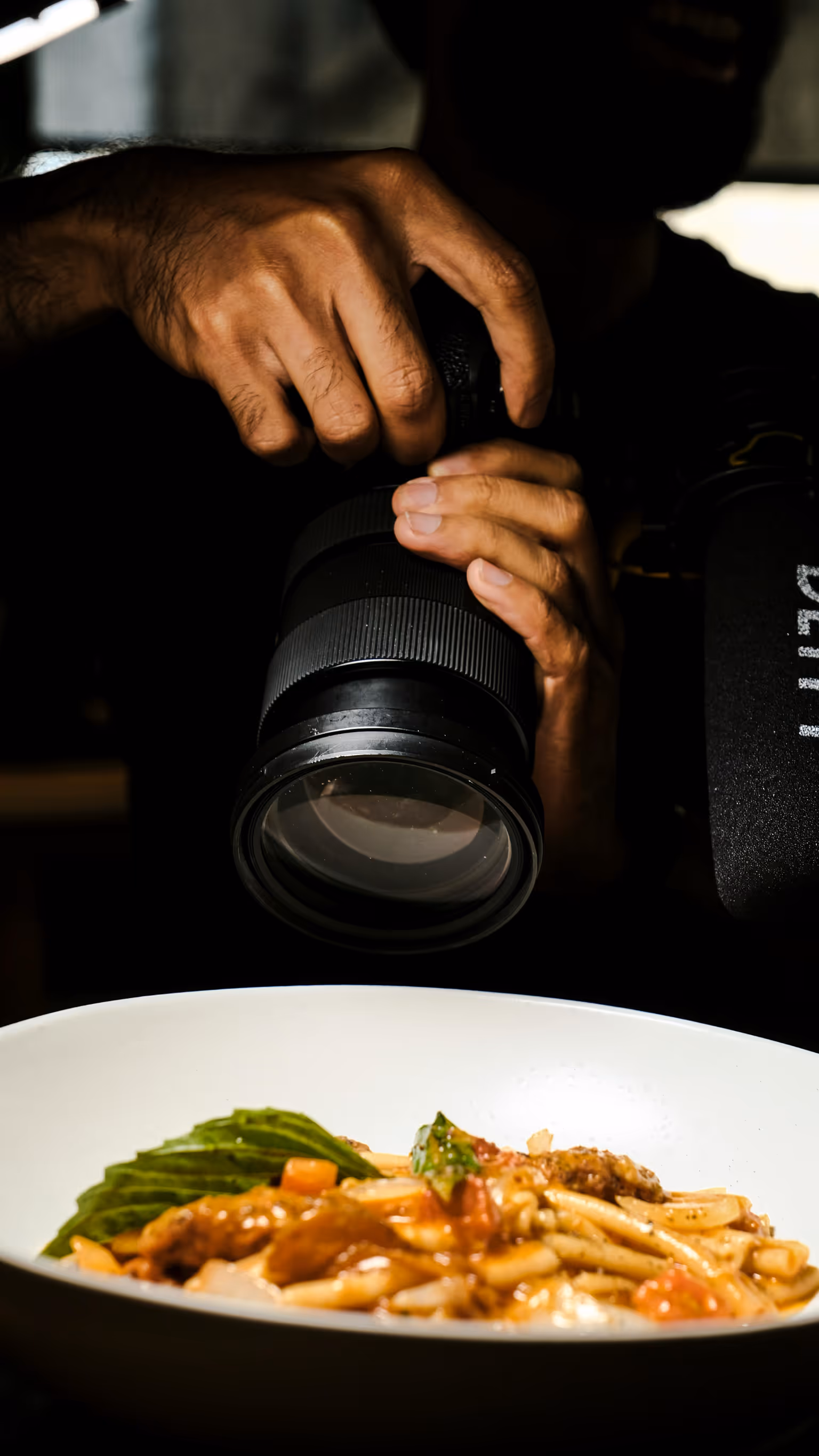 Mano que sostiene una cámara DSLR enfocando un plato con pasta y hojas verdes.