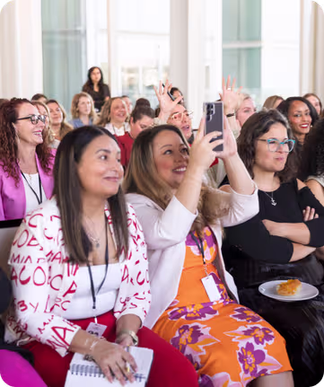 Woman in pink blazer speaking to a group seated around a table with drinks and notebooks in a meeting room.