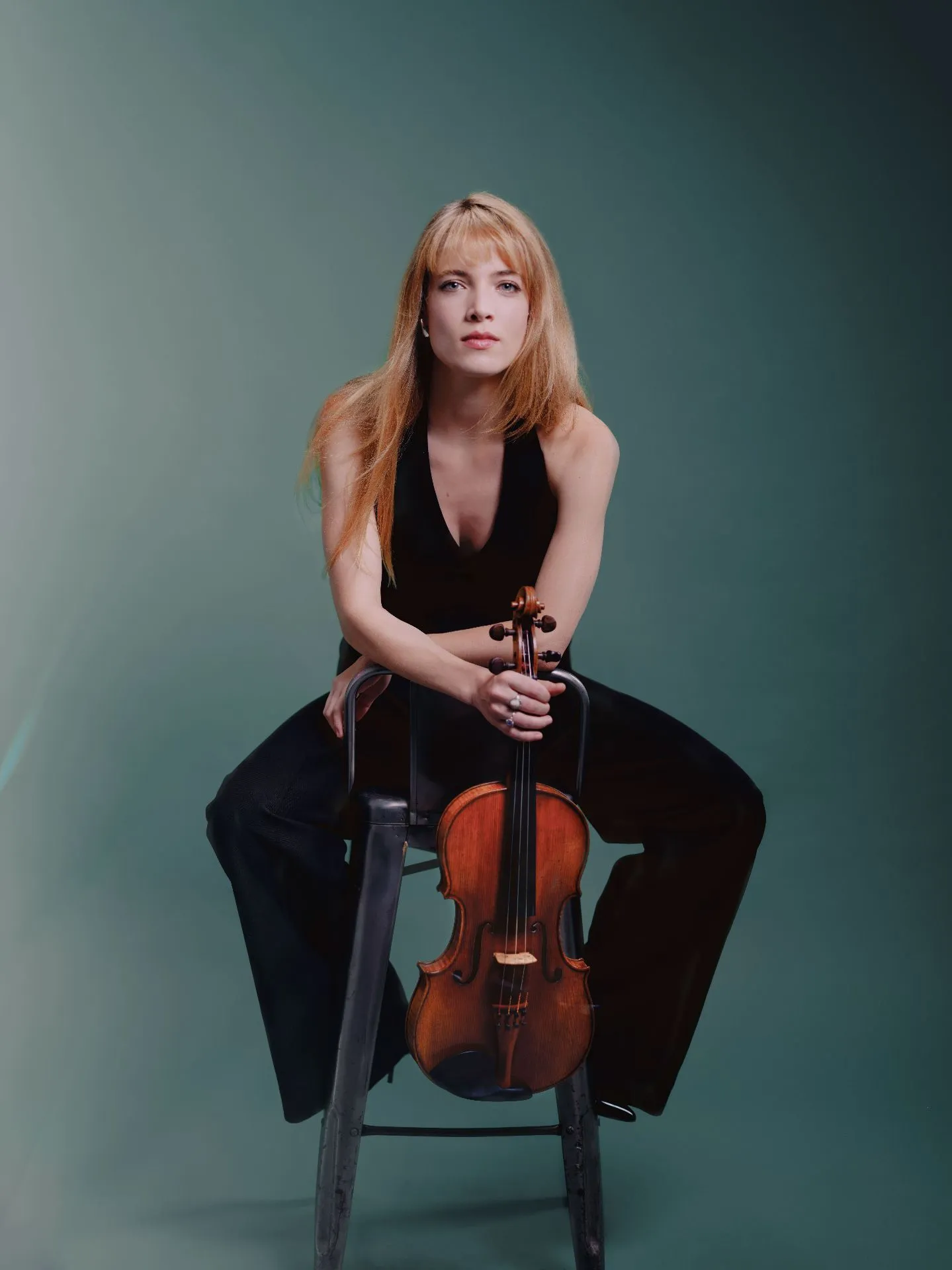 Une femme en tenue noire est assise sur une chaise métallique, tenant un violon verticalement devant elle sur fond vert.