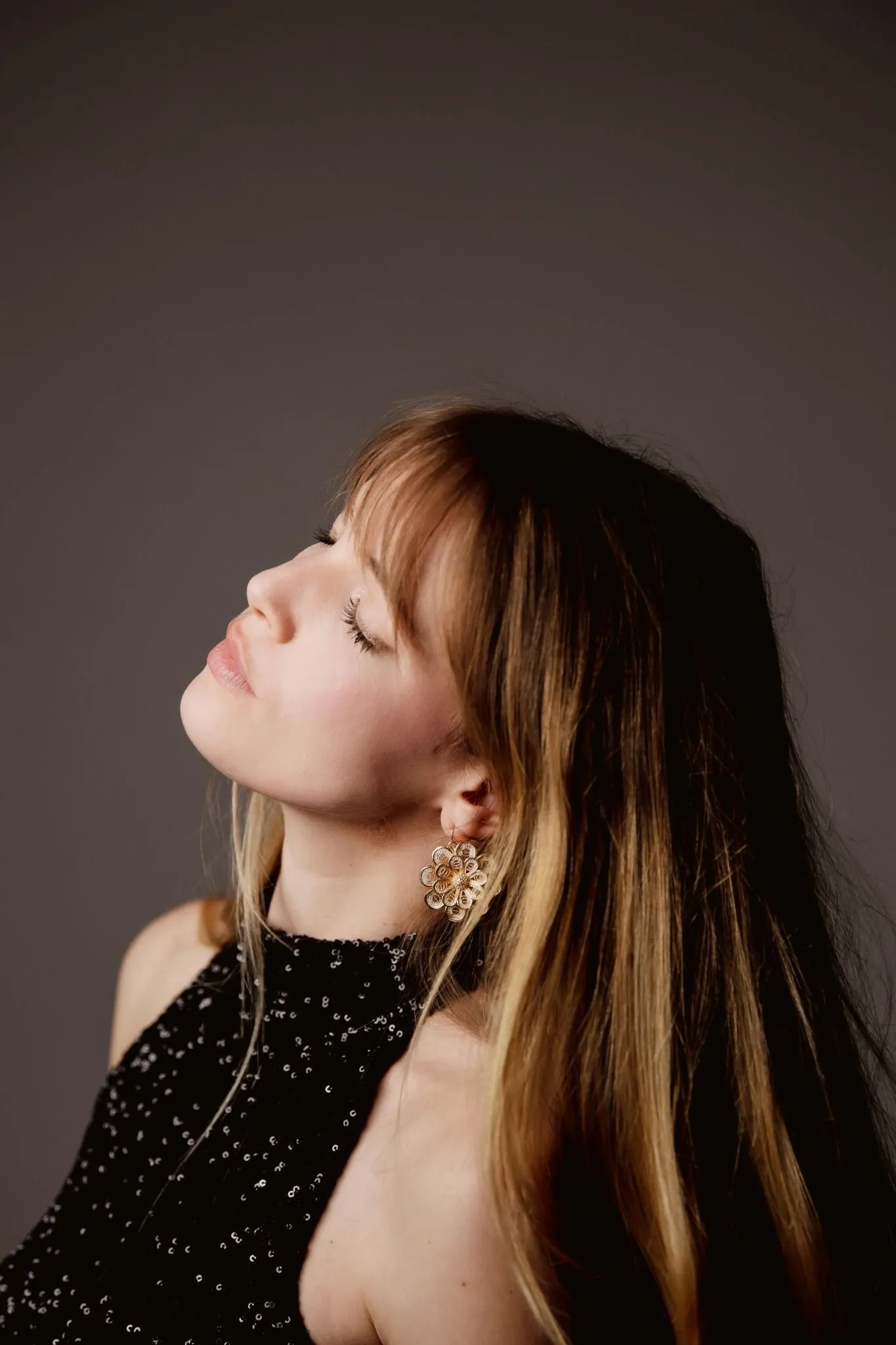 Portrait d'une femme aux cheveux longs blonds regardant vers le haut avec les yeux fermés, portant une robe noire à sequins et une grande boucle d'oreille fleur dorée.