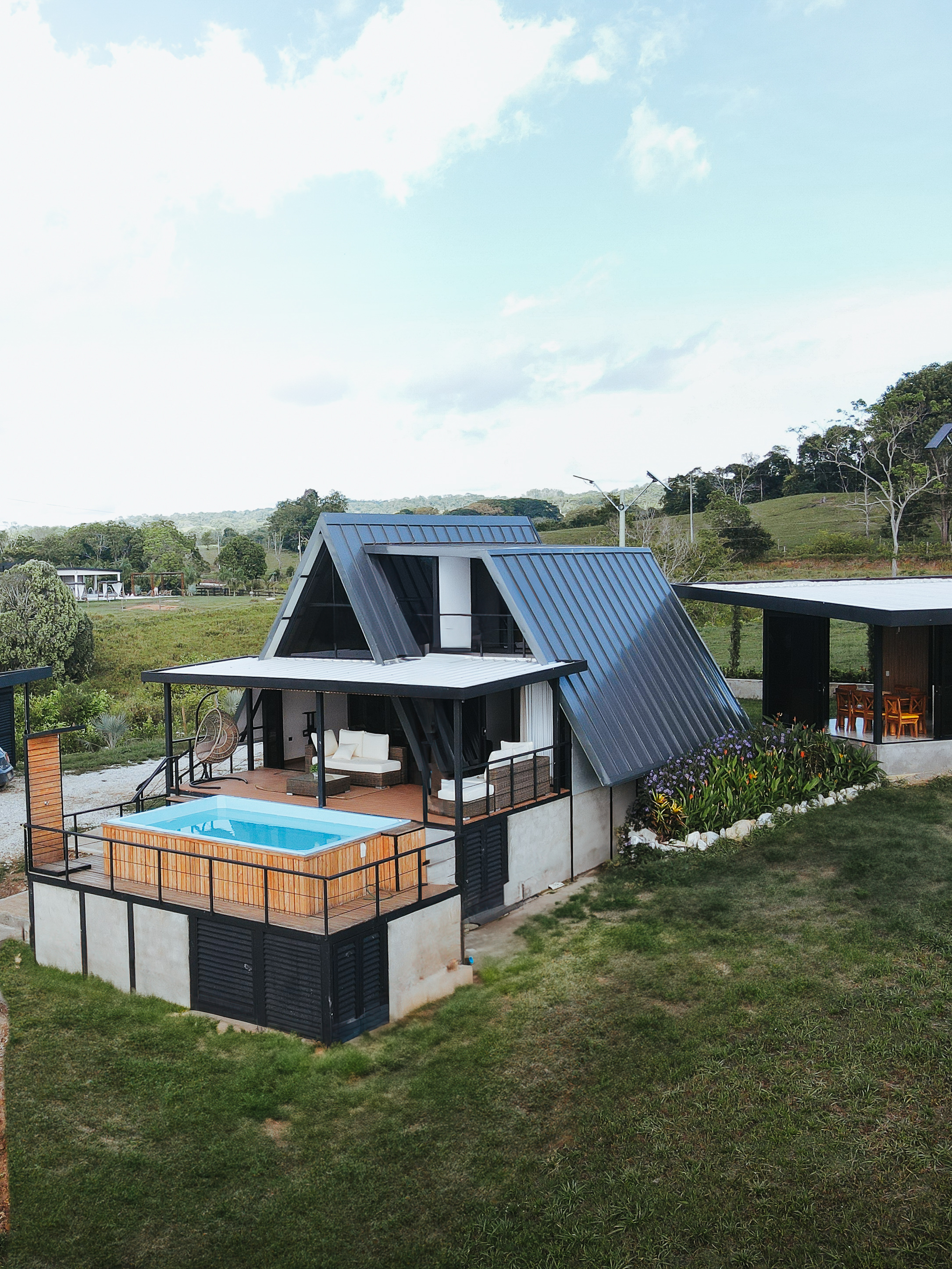 Casa moderna con techo a dos aguas, piscina y terraza amueblada en área rural verde.
