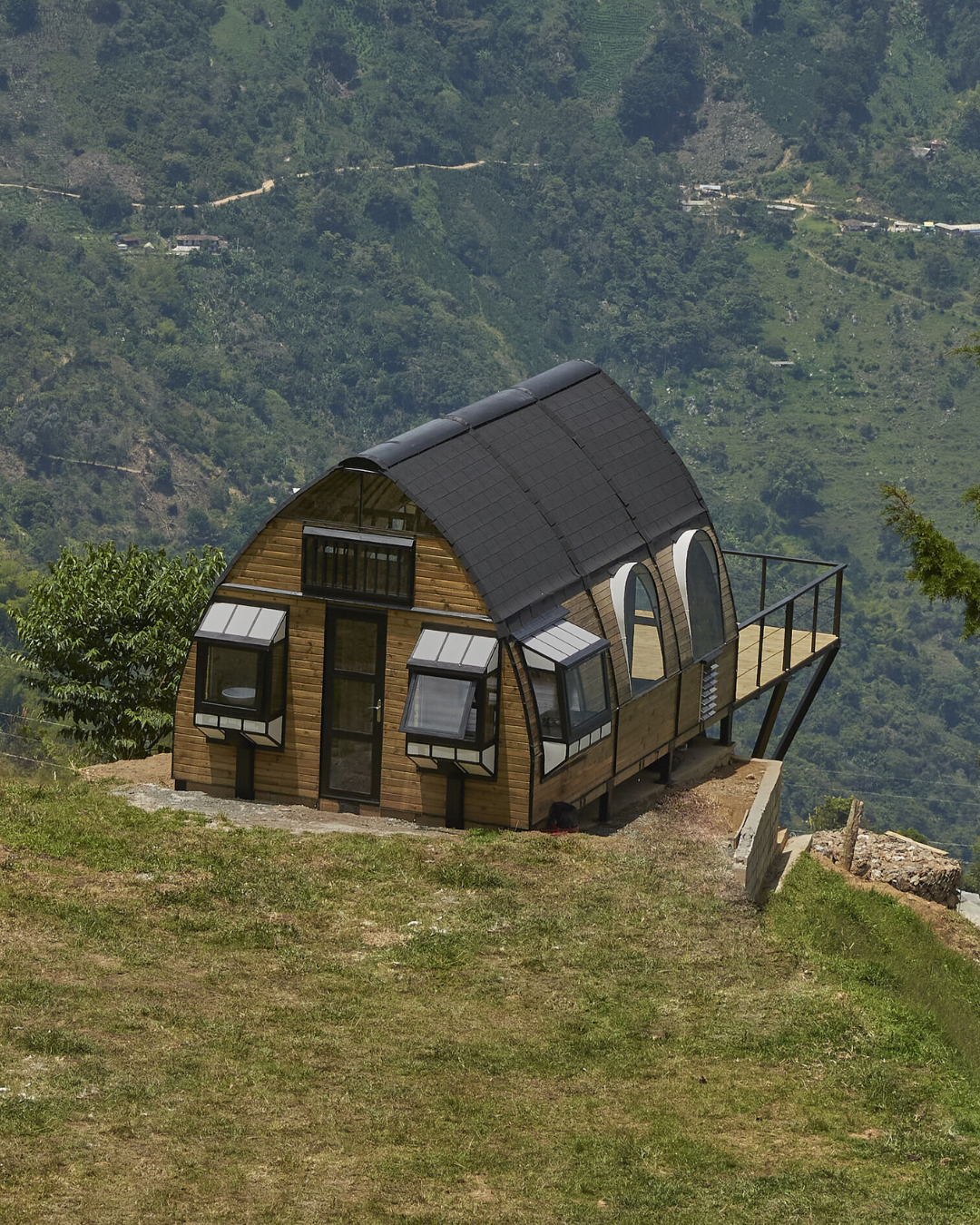 Casa de madera con techo curvo negro y ventanas amplias en ladera con vegetación de fondo.