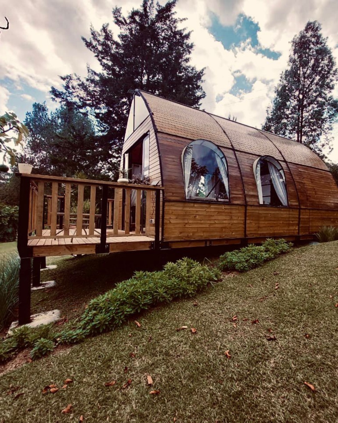 Cabaña de madera elevada con ventanas curvas y terraza en jardín verde con árboles altos.