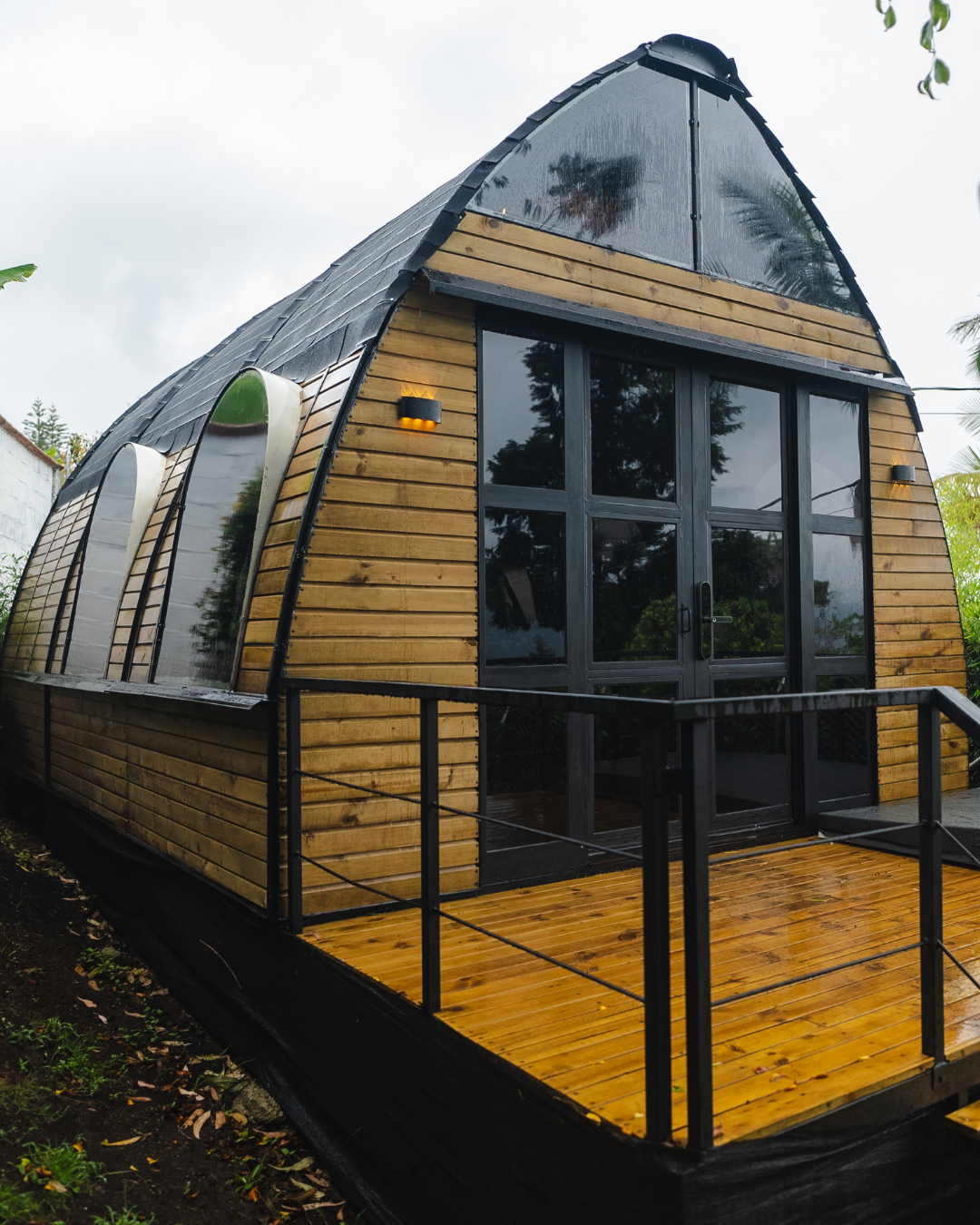 Casa moderna de madera con techo curvado negro y ventanas grandes reflectantes en un día lluvioso.