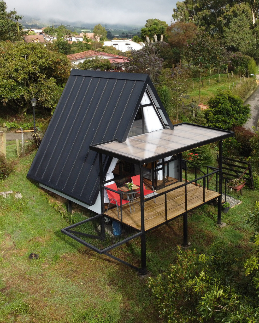 Cabaña tipo A con terraza cubierta, ubicada en un área verde con sillas rojas y mesa pequeña.