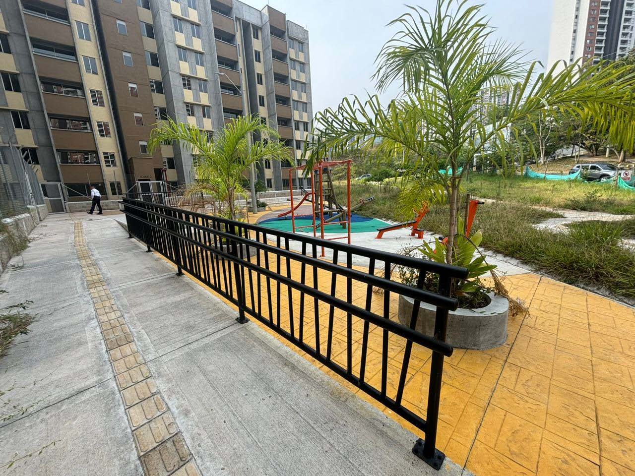 Área con rampa accesible, plantas, juegos infantiles y edificio residencial al fondo.