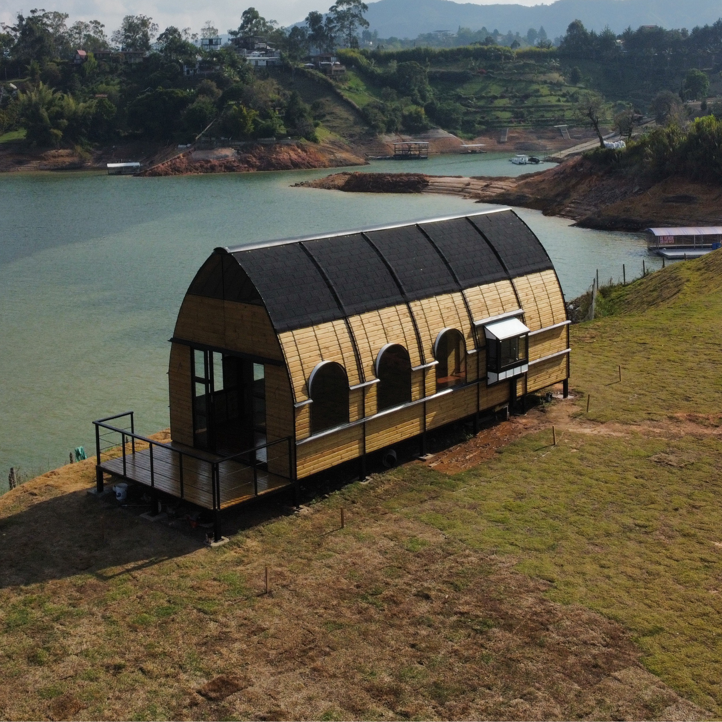 Casa de madera con techo curvado junto a un lago en un paisaje montañoso y verde.