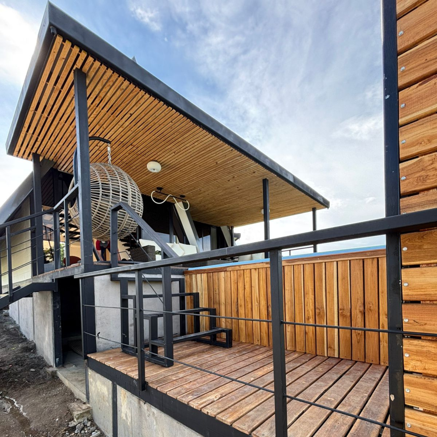 Terraza de madera en casa moderna con barandal negro, techo de listones y silla colgante de mimbre.
