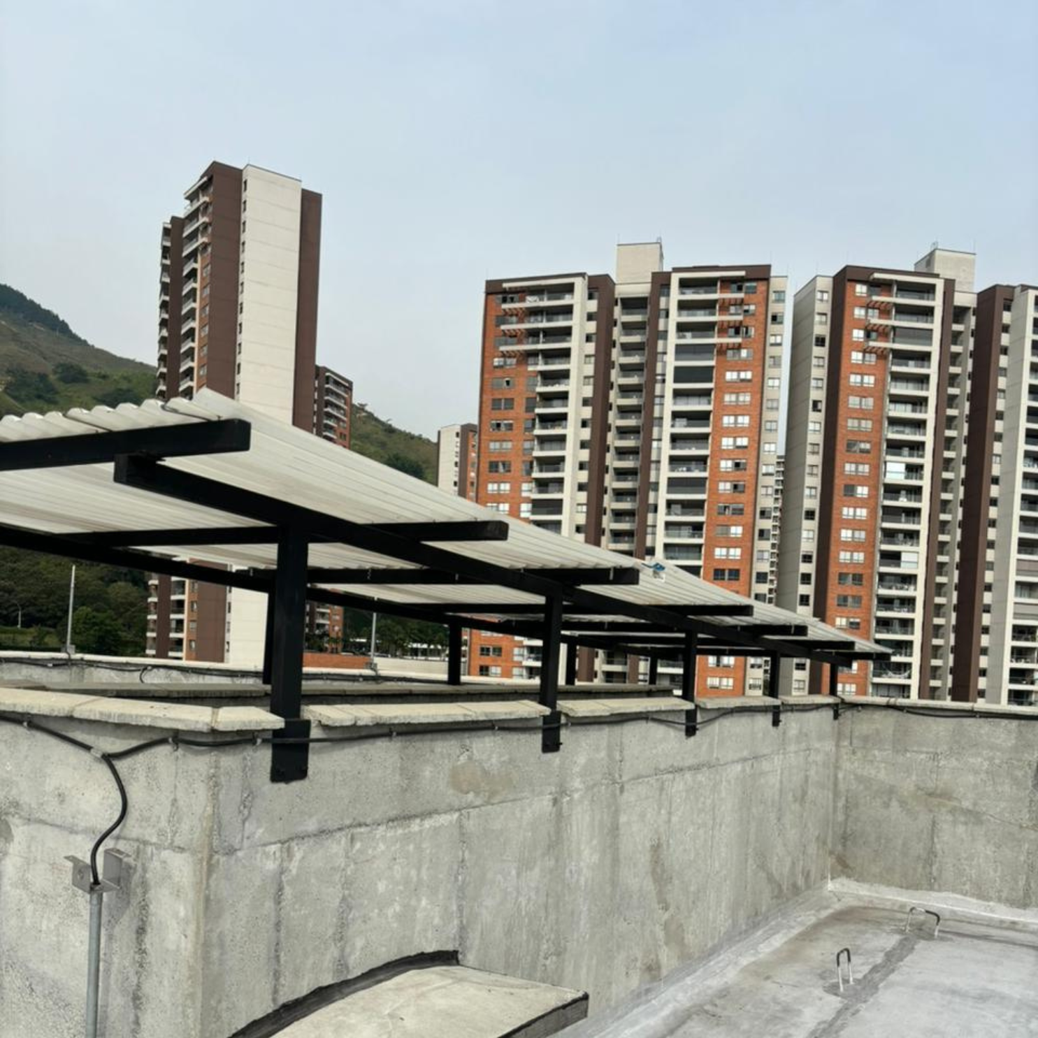 Estructura metálica con paneles sobre muro de concreto, edificios residenciales y colina al fondo.