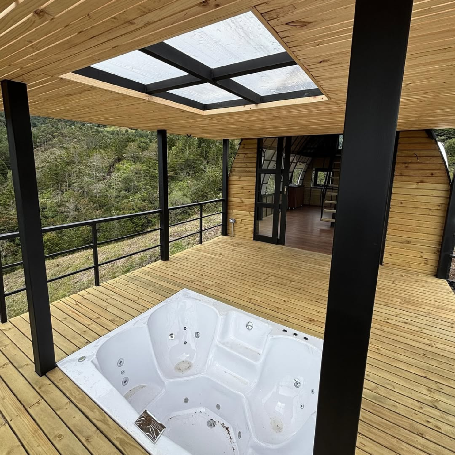 Jacuzzi blanco empotrado en terraza de madera con techo de paneles transparentes y vista a bosque.