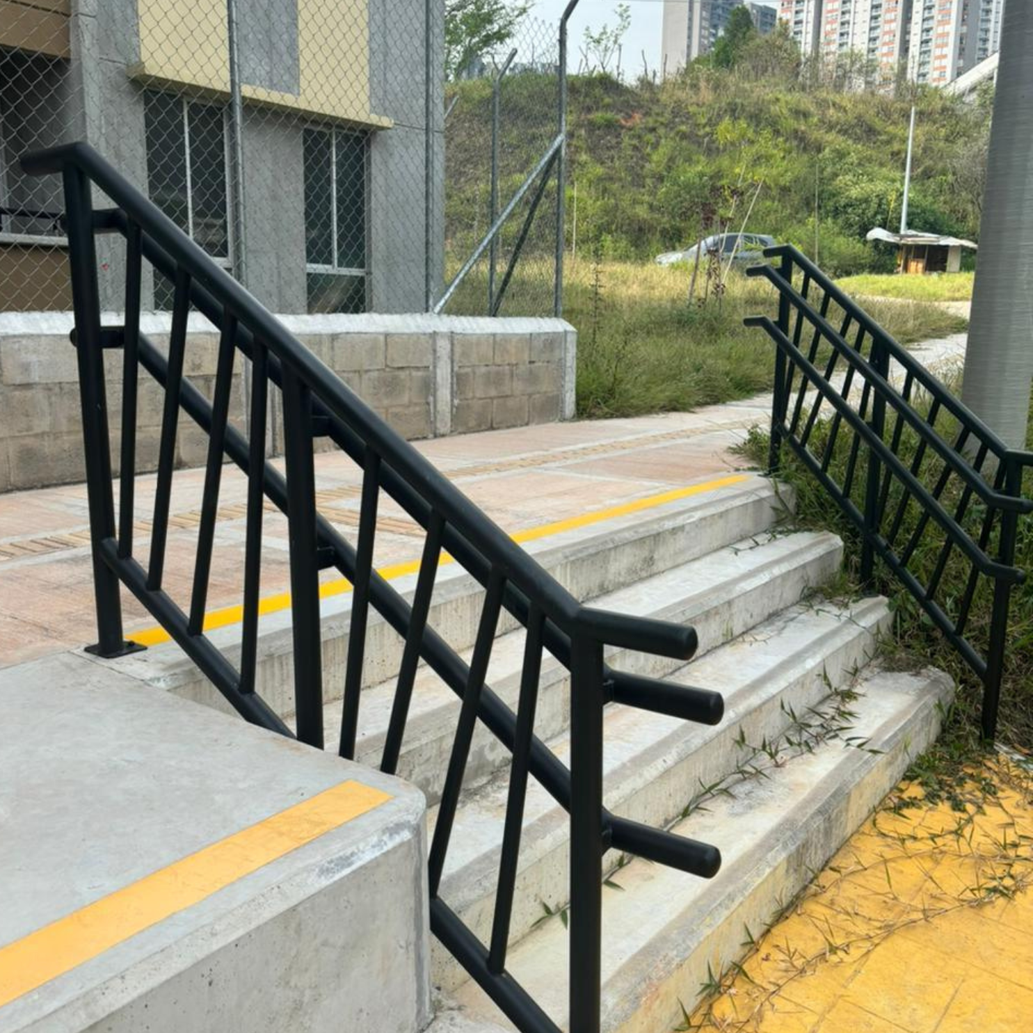 Escaleras de concreto con barandas negras y maleza creciendo entre los escalones al aire libre.