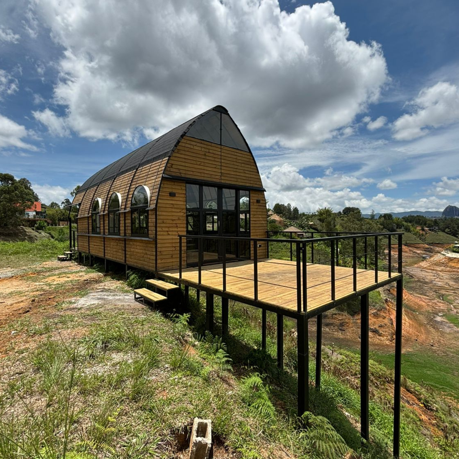 Casa de madera elevada con terraza, ventanales arqueados y cielo nublado en zona rural verde.