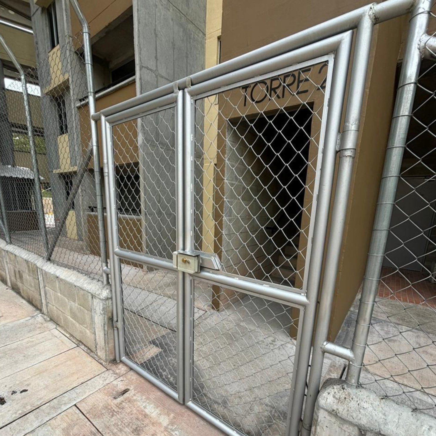 Portón de malla metálica con candado frente a edificio con letrero que dice TORRE 2.